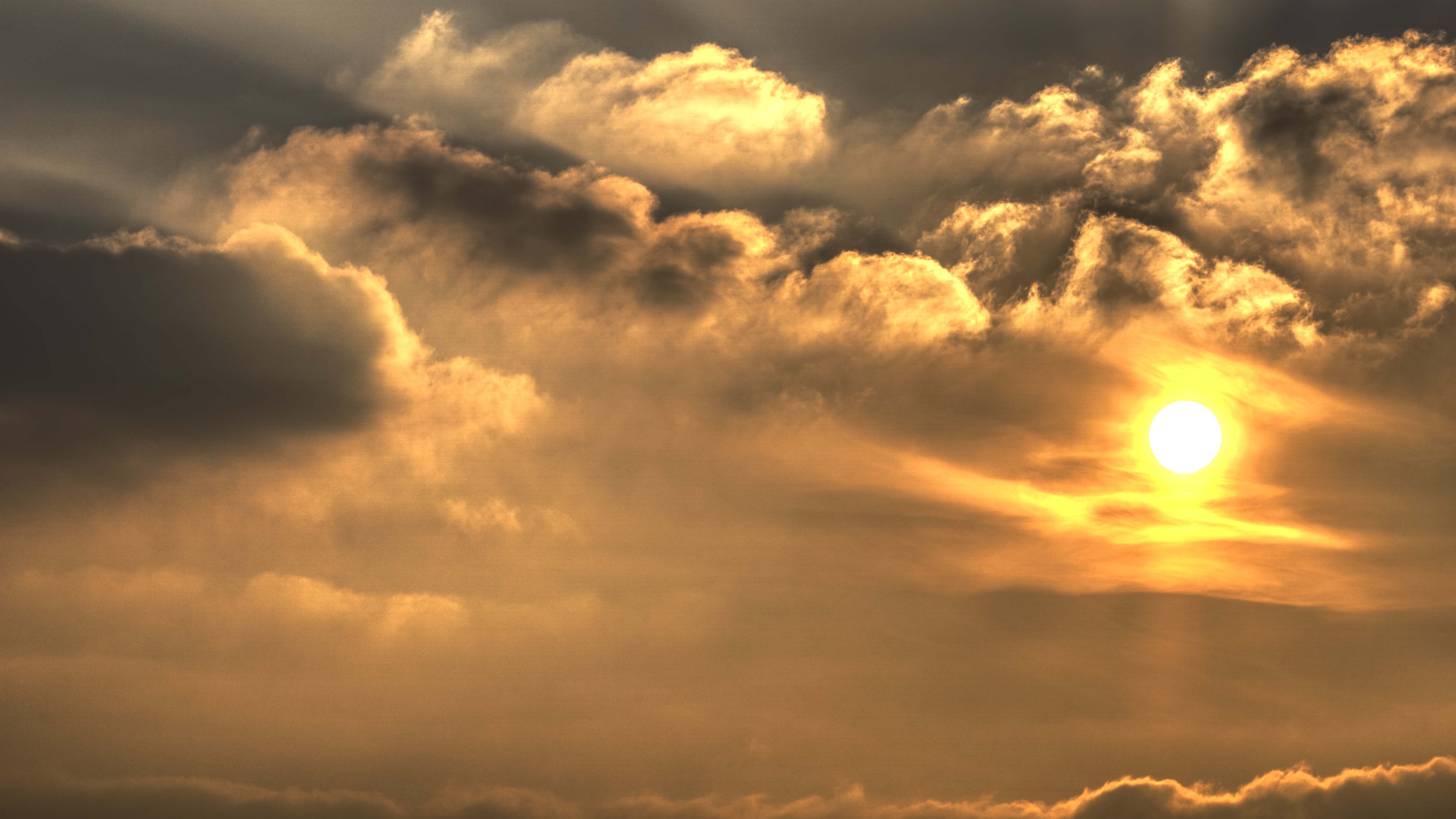 Sonne zwischen dichten, dunkelgrauen Wolken, deren Ränder vom goldenen Sonnenlicht beleuchtet werden.
