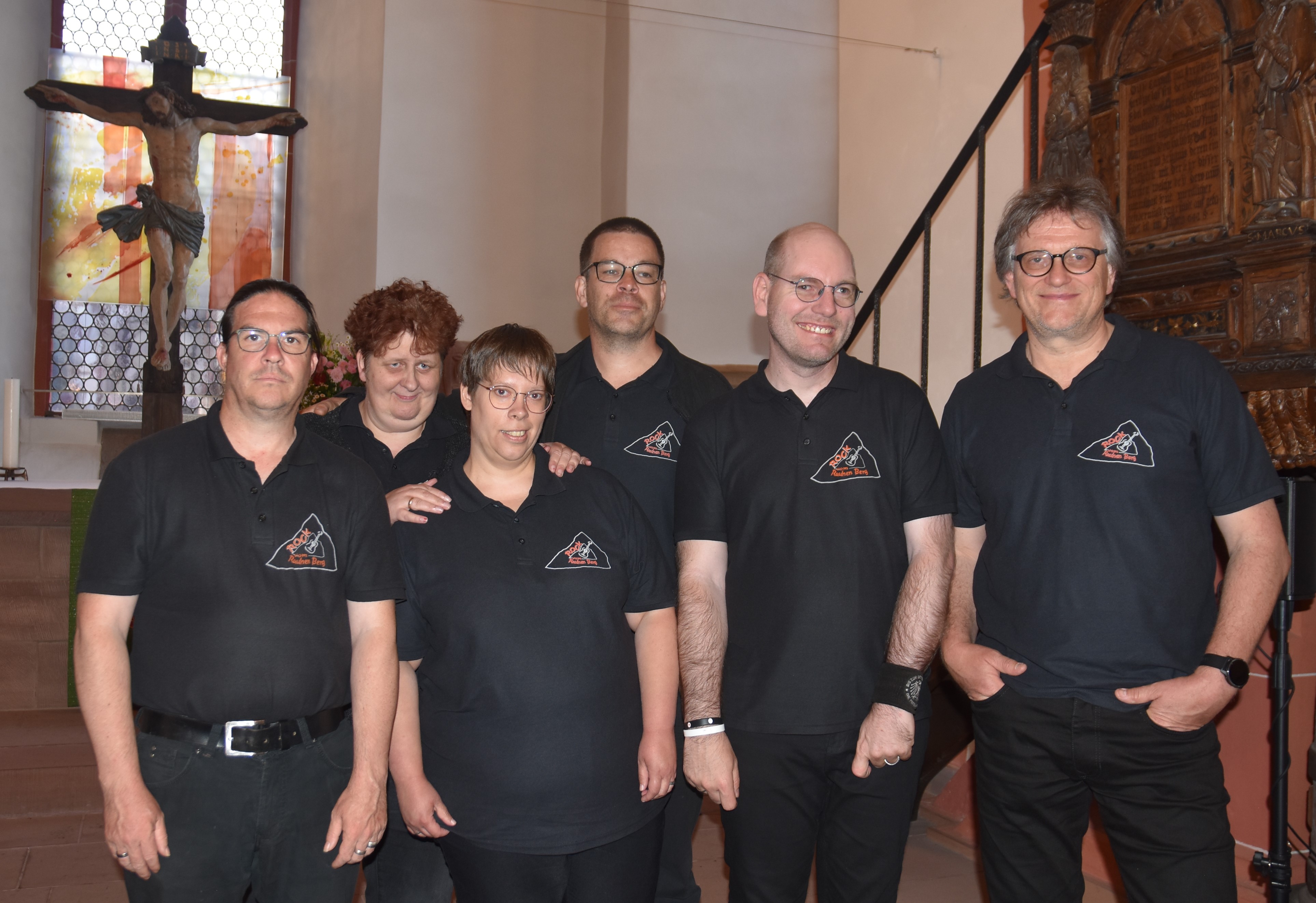 Gruppenfoto der Mitglieder der inklusiven Band "Rocker vom Rauhen Berg" mit ihrem Bandleader Thomas Appel. Alle tragen schwarze T-Shirts und stehen im Altarraum der Niddaer Kirche.