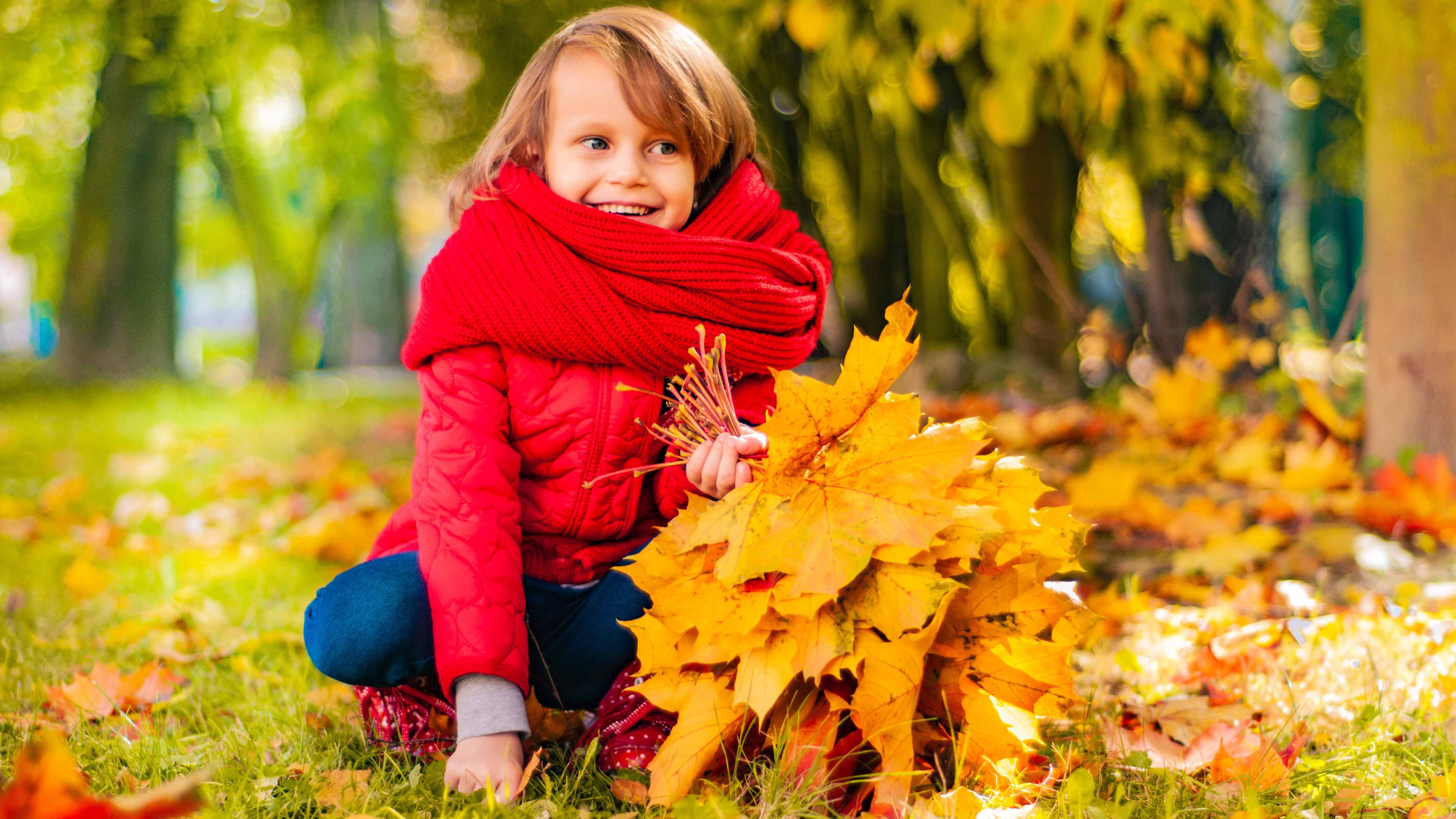 Ein lächelndes Kind mit roter Jacke und rotem Schal hockt im Freien und hält einen großen Strauß gelber Herbstblätter in der Hand.