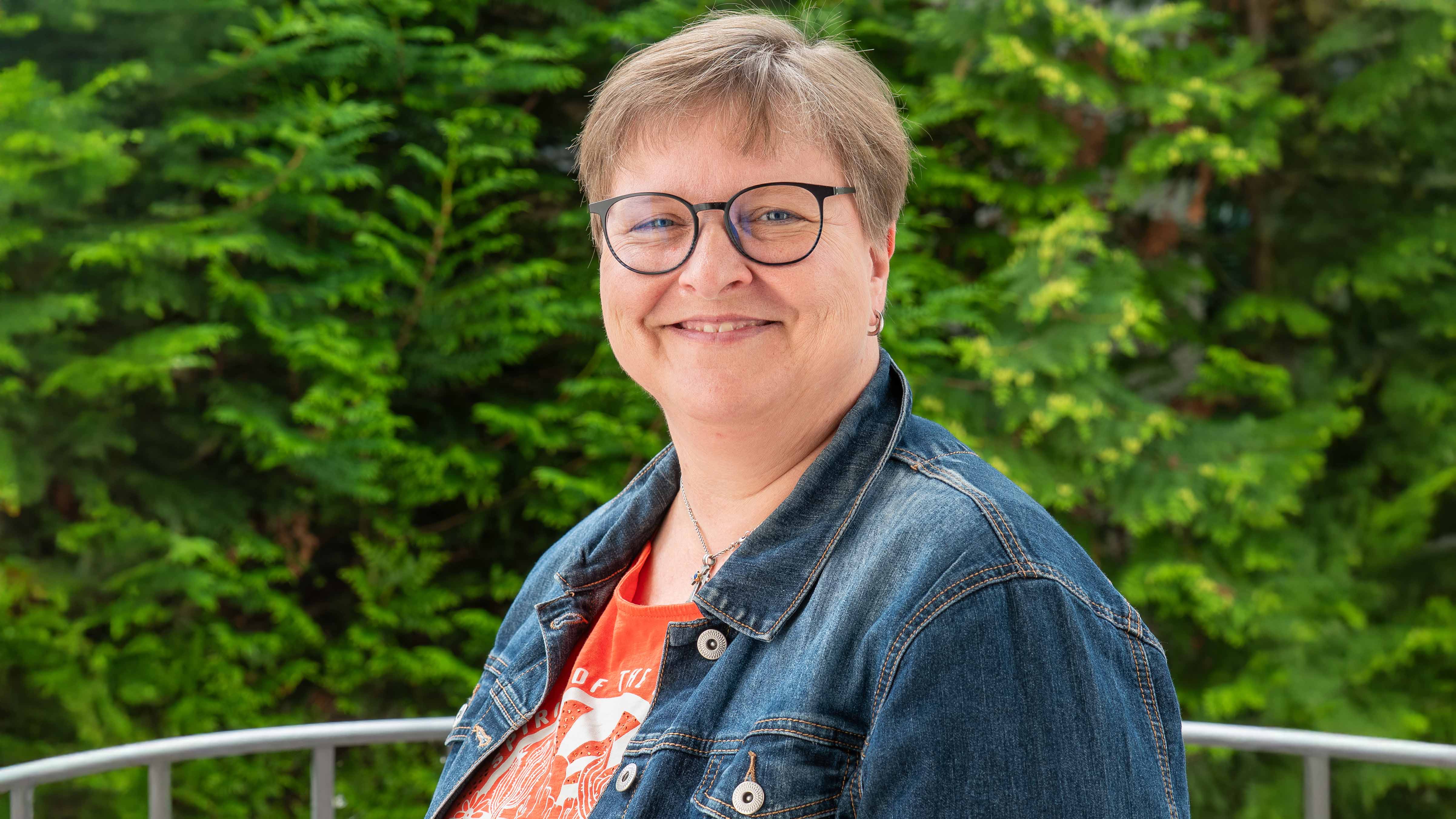 Anke Thürigen lächelt in die Kamera. Sie trägt eine dunkle Brille, eine blaue Jeansjacke und ein orangefarbenes T-Shirt. Im Hintergrund sind dichte grüne Sträucher zu sehen.