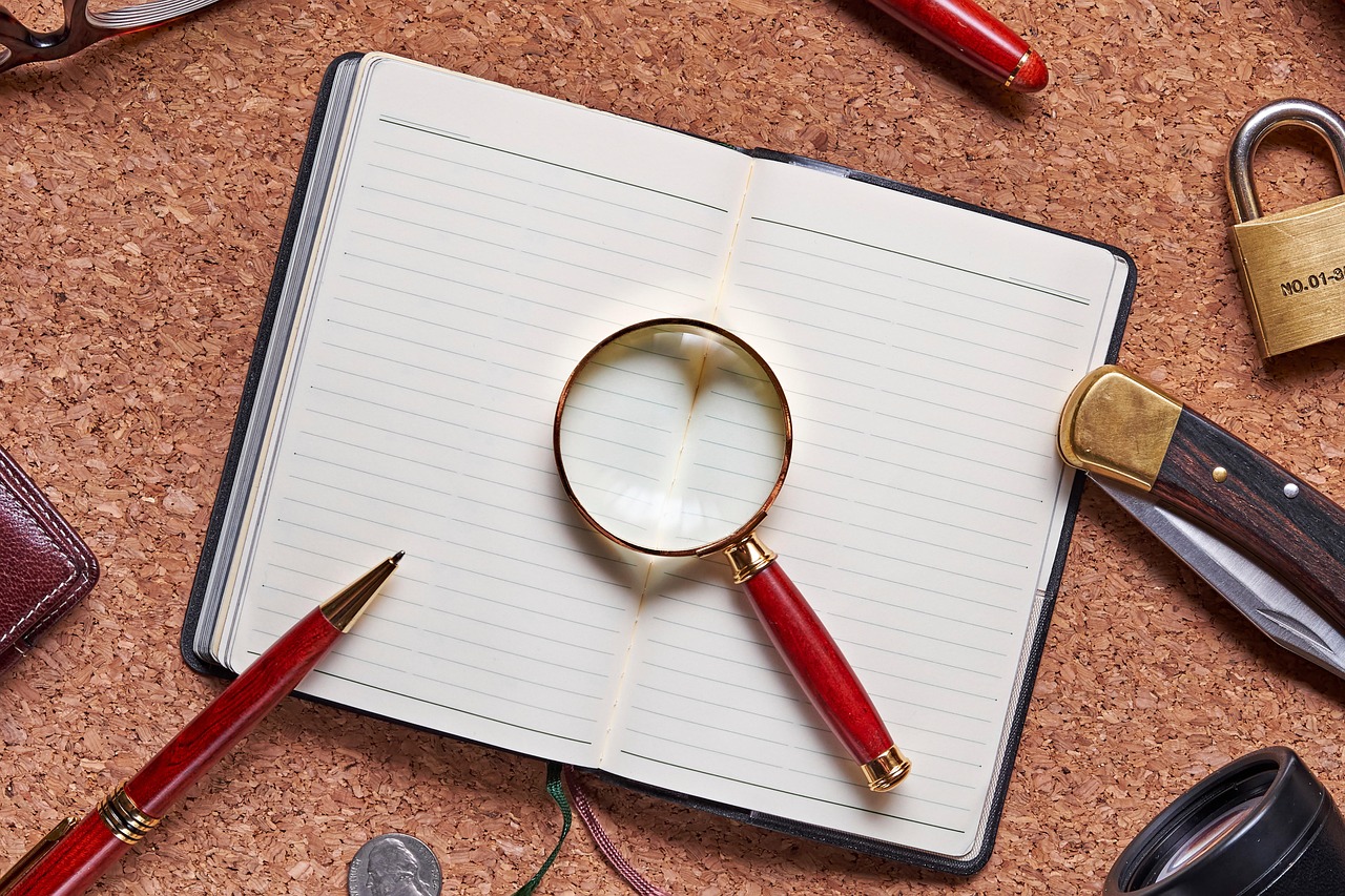 Auf einem Tisch liegt ein leeres Buch mit einer Lupe, einem Stift, einem Taschenmesser und einem Vorhängeschloss
