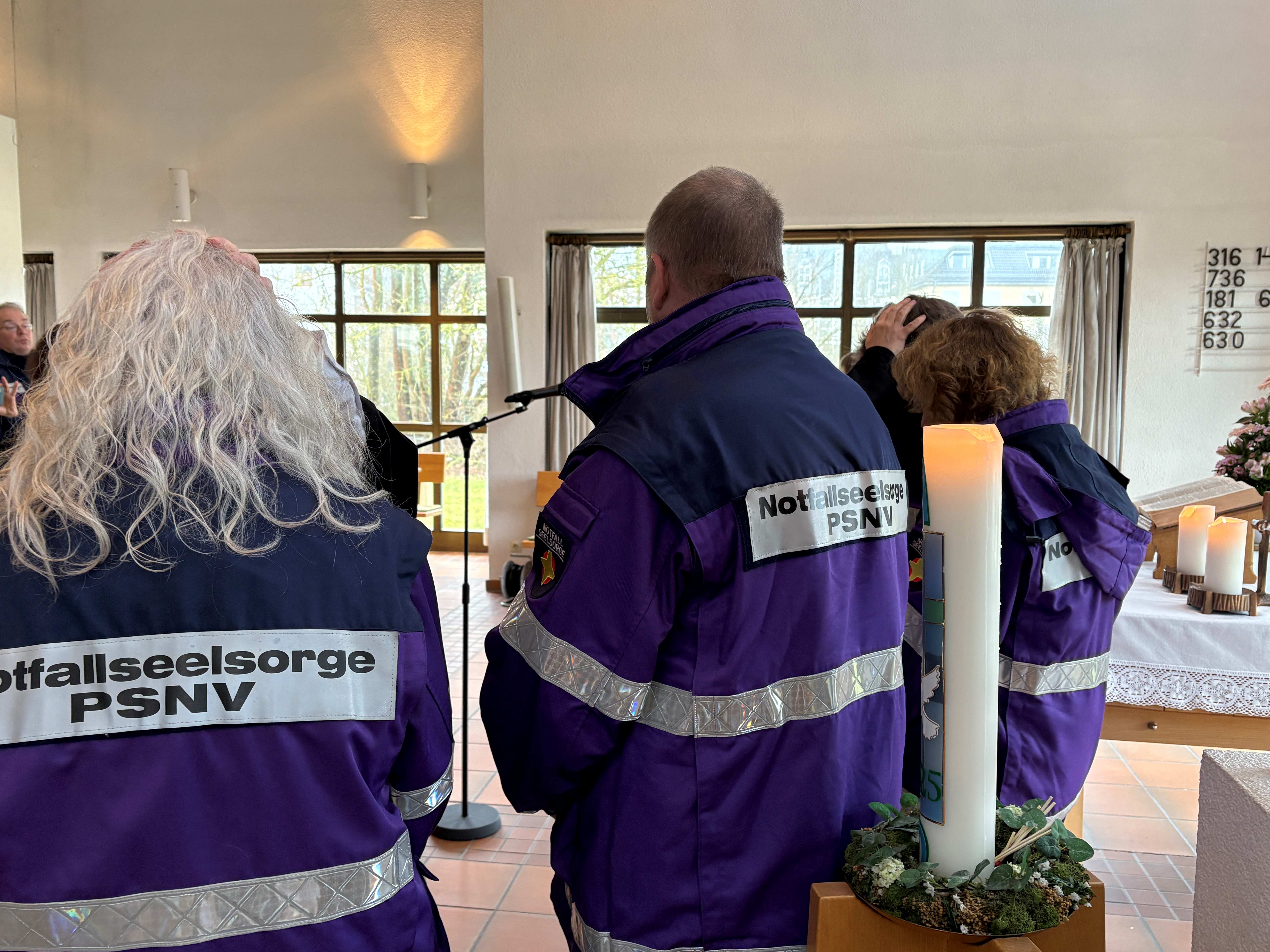 Menschen in violetten Jacken auf denen "Notfallseelsorge PSNV" drauf steht, stehen im Altarraum vor einer Kerze und dem Altar und werden durch eine Frau gesegnet, die ihre Hände auf den Kopf einer Frau legt.