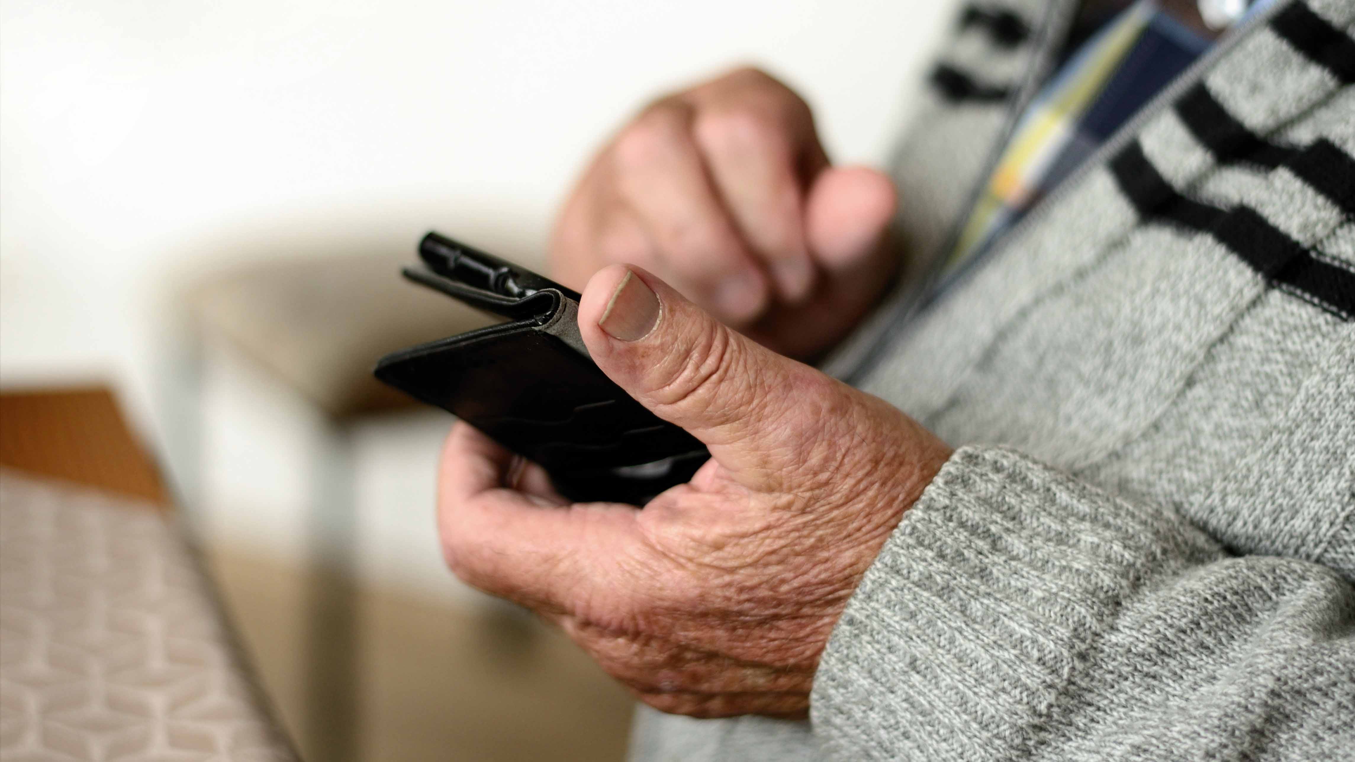 Smartphone_Senior_C_congerdesign_Pixabay_16zu9.jpg Ältere Hände halten ein schwarzes Smartphone; ein Daumen liegt auf dem Bildschirm.