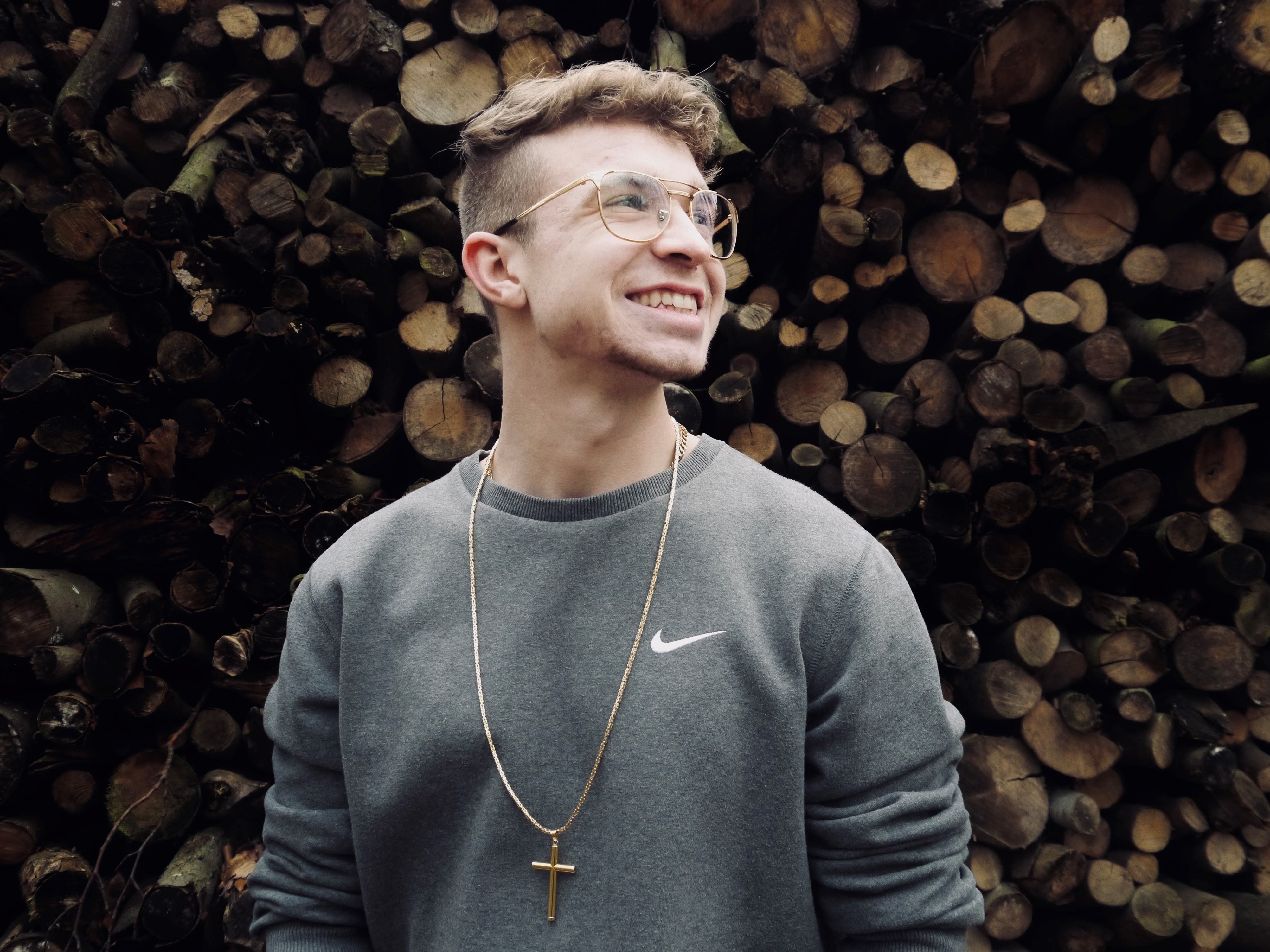 Colin McLemore, dunkelblonde Haare und Brille, trägt um den Hals eine Kette mit einem großen goldenen Kreuz. Er steht vor einem Holzstapel und richtet seinen Blick zum Himmel.